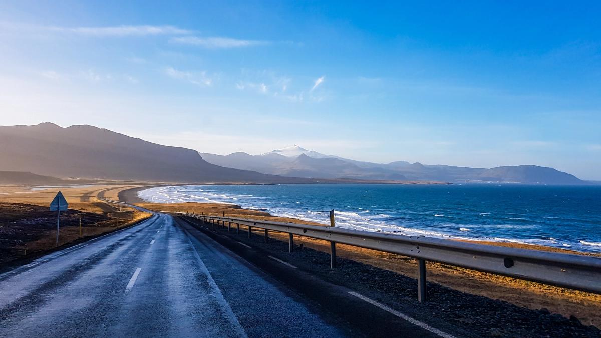 A wet road curves along an ocean coast with distant mountains.