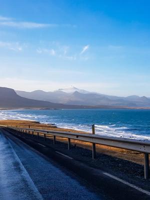 A wet road winds along a rugged coastline with a blue ocean and distant mountains under a clear sky.