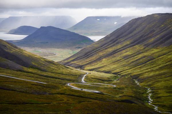 A winding road and stream descend through a vast green mountain valley towards a distant lake under a cloudy sky.
