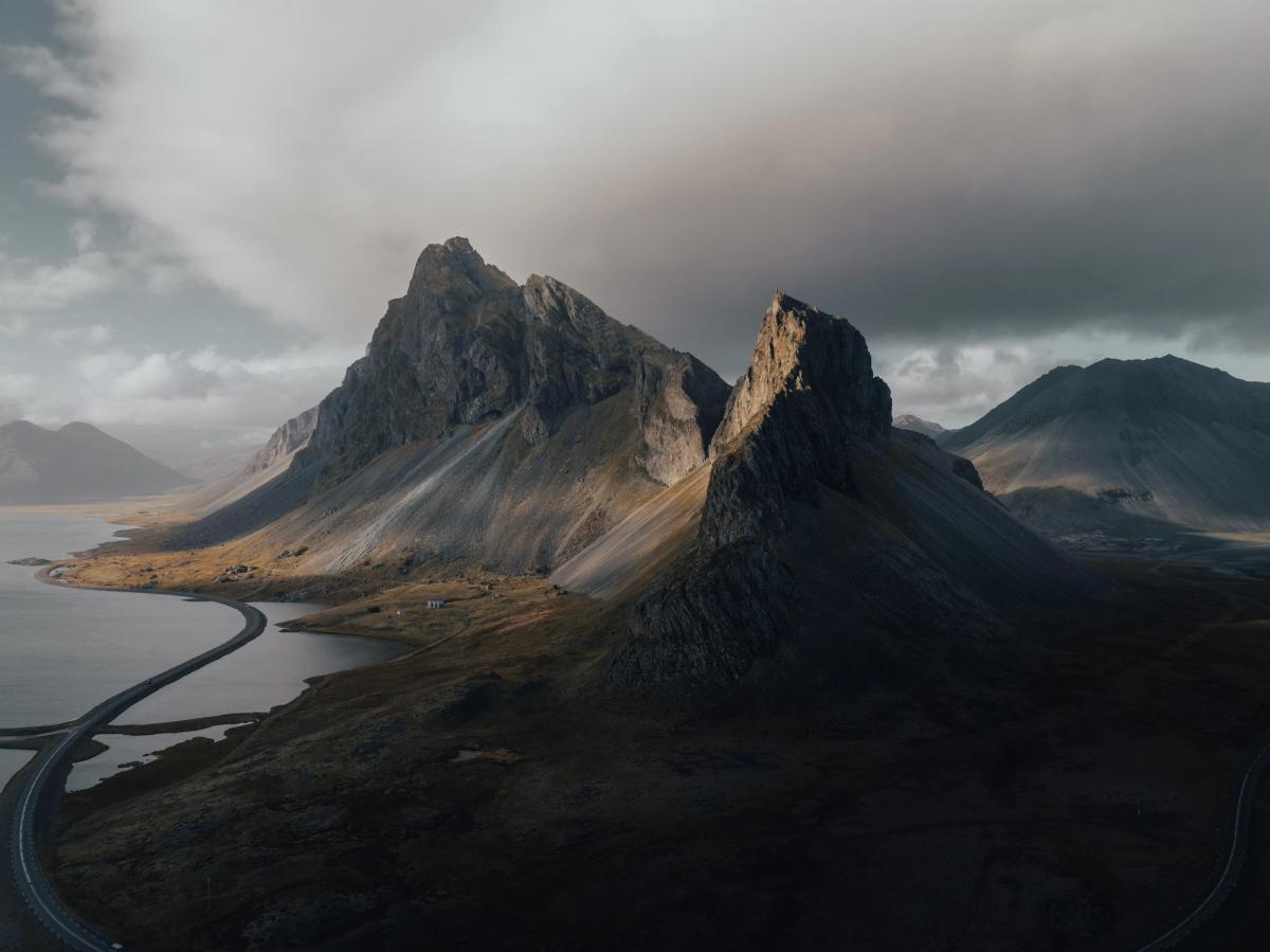 Majestic jagged mountains overlook a curving road and water, with sunlight breaking through dramatic cloudy skies.