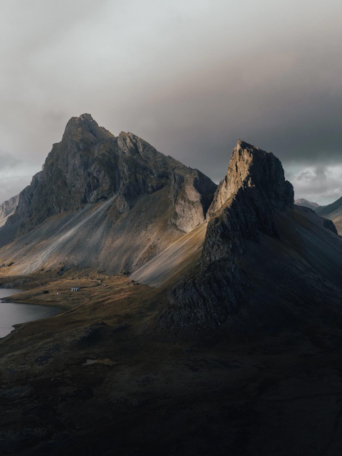 Majestic jagged mountains overlook a curving road and water, with sunlight breaking through dramatic cloudy skies.