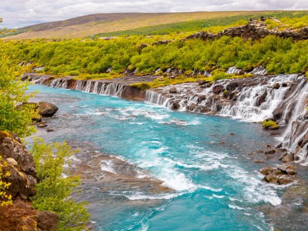 Hraunfossar Waterfalls with green grass and trees around it.