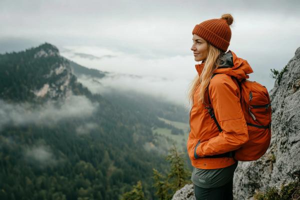 Mujer con chaqueta y gorro naranja, mochila a la espalda, contemplando un paisaje montañoso boscoso envuelto en niebla.