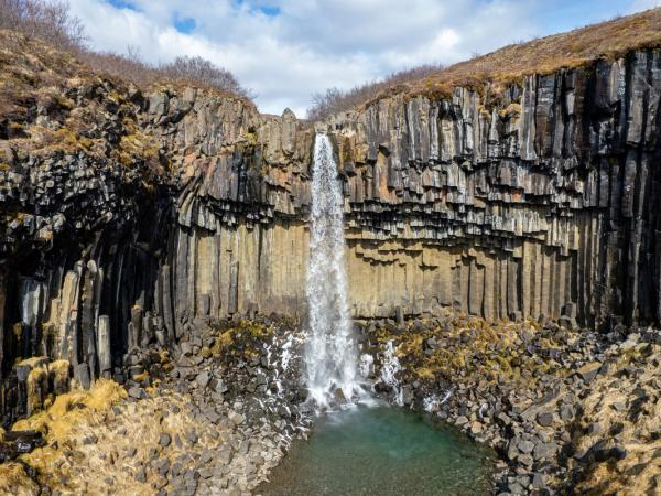 Waterfall plunging over dark, geometric basalt columns into a clear pool.