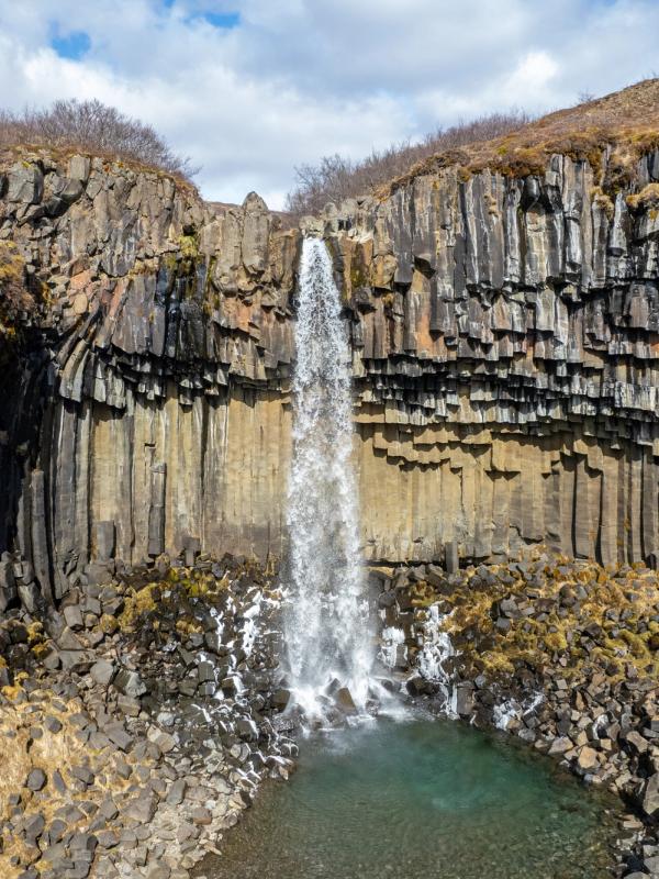 Waterfall plunging over dark, geometric basalt columns into a clear pool.