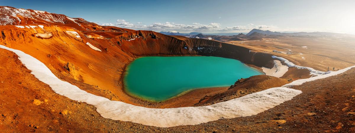 Panoramic view of a vibrant turquoise crater lake surrounded by reddish-brown volcanic terrain with patches of snow.
