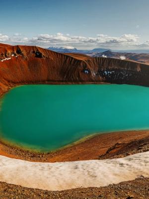 Panoramic view of a vibrant turquoise crater lake surrounded by reddish-brown volcanic terrain with patches of snow.