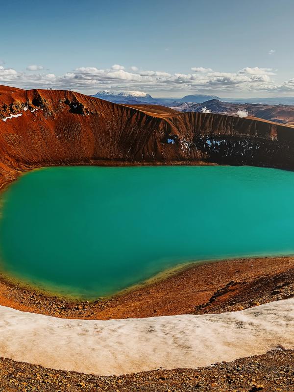 Panoramic view of a vibrant turquoise crater lake surrounded by reddish-brown volcanic terrain with patches of snow.
