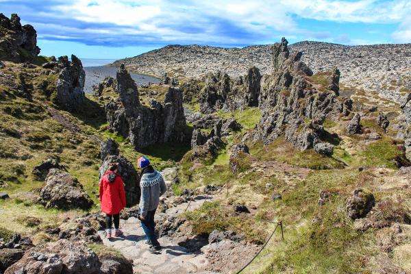 Two people walk on a path through a rugged volcanic landscape with tall rock formations, leading towards a black sand beach and the ocean.