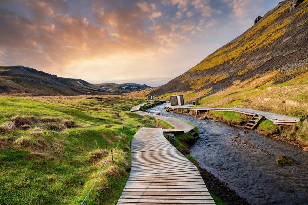 A winding wooden boardwalk follows a river through a green valley with a steep, streaked hill under an orange sunset sky.
