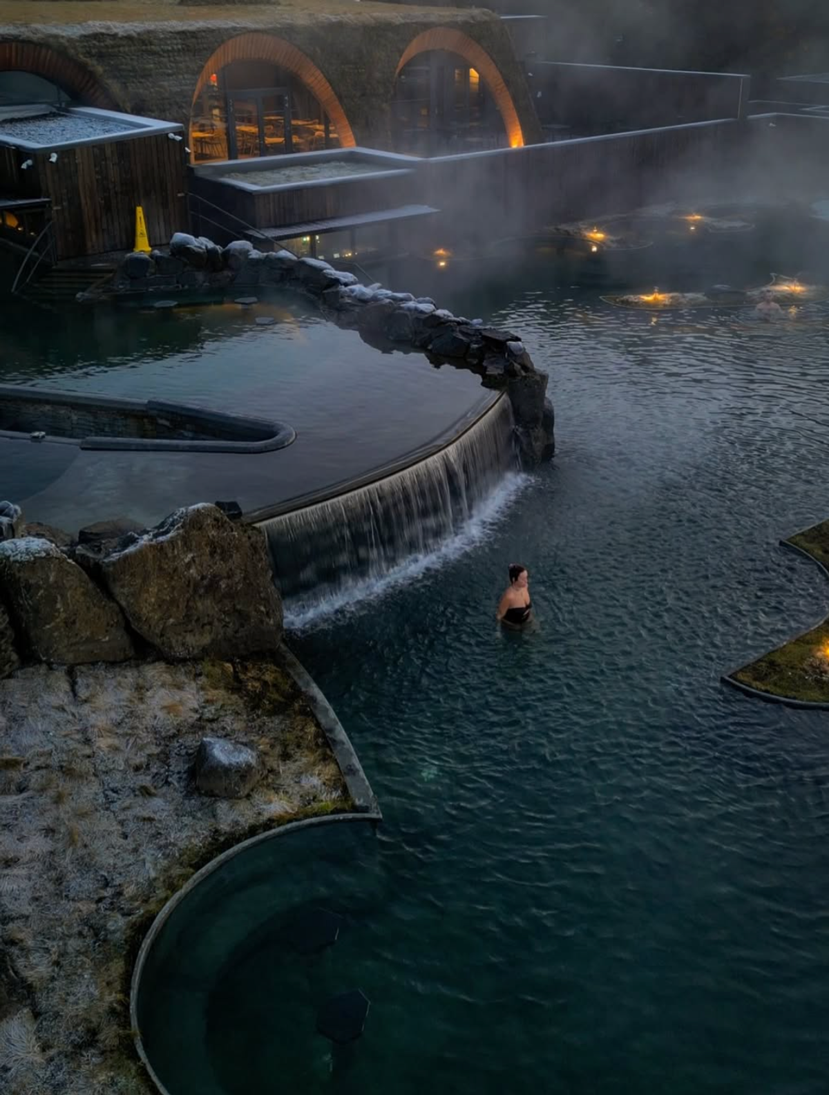 Una persona nada en una piscina geotérmica al aire libre, envuelta en niebla y con una cascada, junto a edificios semienterrados en la tierra al anochecer.