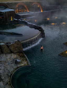 Una persona nada en una piscina geotérmica al aire libre, envuelta en niebla y con una cascada, junto a edificios semienterrados en la tierra al anochecer.