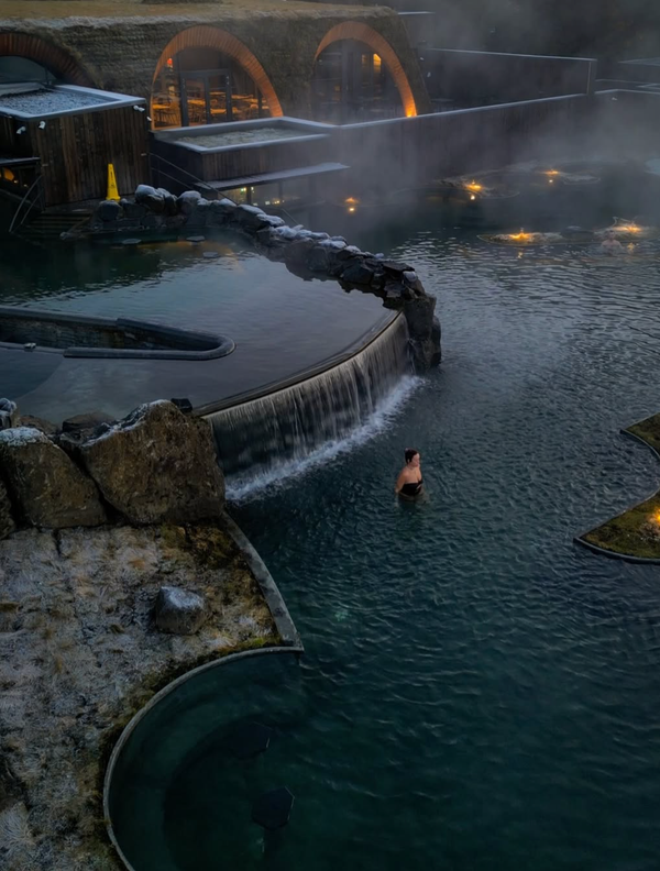 A woman in a steaming outdoor geothermal pool with a waterfall, frosted rocks, and a distinctive grass-roofed building.