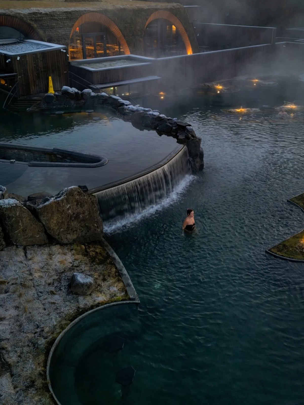 Una persona nada en una piscina geotérmica al aire libre, envuelta en niebla y con una cascada, junto a edificios semienterrados en la tierra al anochecer.