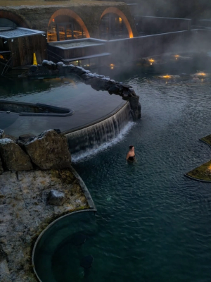 Una mujer en una piscina geotérmica al aire libre, llena de vapor, con una cascada, rocas heladas y un edificio característico con techo de hierba.