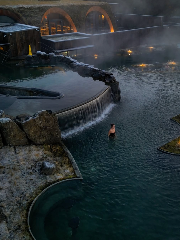 A woman in a steaming outdoor geothermal pool with a waterfall, frosted rocks, and a distinctive grass-roofed building.