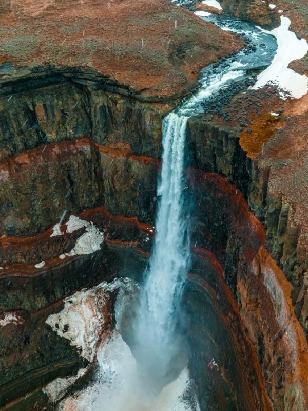 A powerful waterfall plunges into a rocky canyon with reddish-brown terrain and patches of snow.