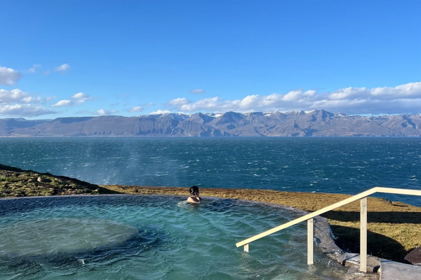 Person relaxing in a geothermal pool overlooking a fjord with distant snowy mountains under a blue sky.