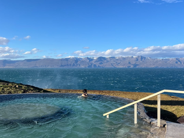 A person relaxes in a steaming outdoor geothermal pool overlooking a windy ocean and snow-capped mountains under a blue sky.
