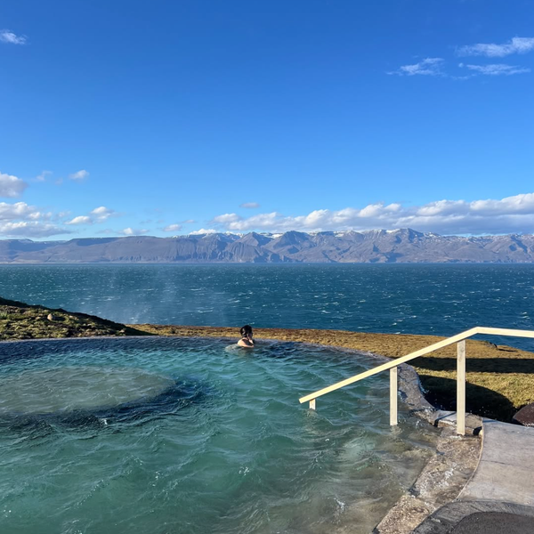 A person in a steamy geothermal pool overlooking a vast body of water and distant mountains under a blue sky.