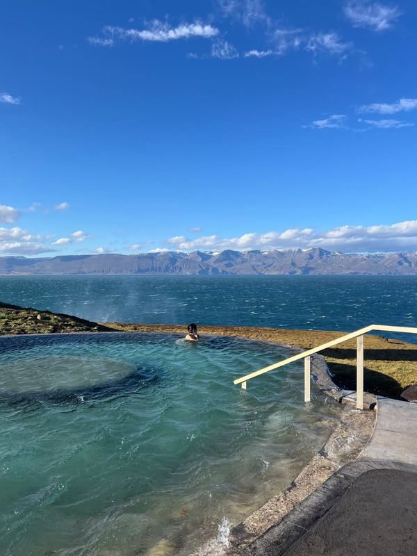 A person in an outdoor geothermal pool overlooking a bay with mountains under a blue sky.