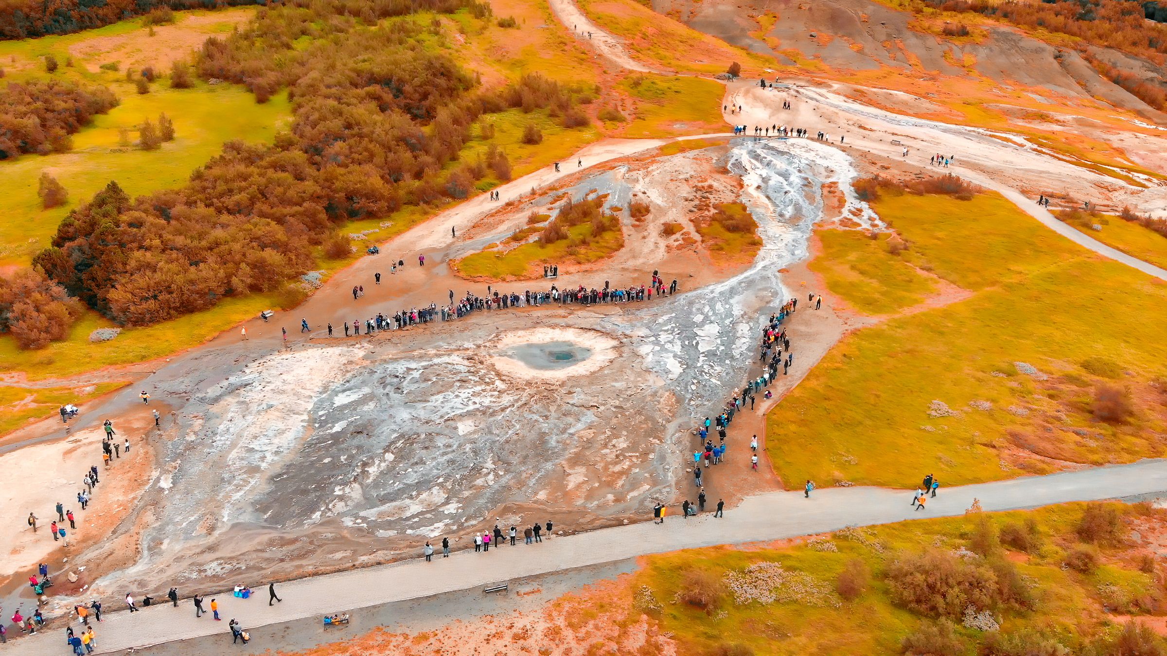 Aerial view of tourists around a geyser in a golden geothermal landscape.