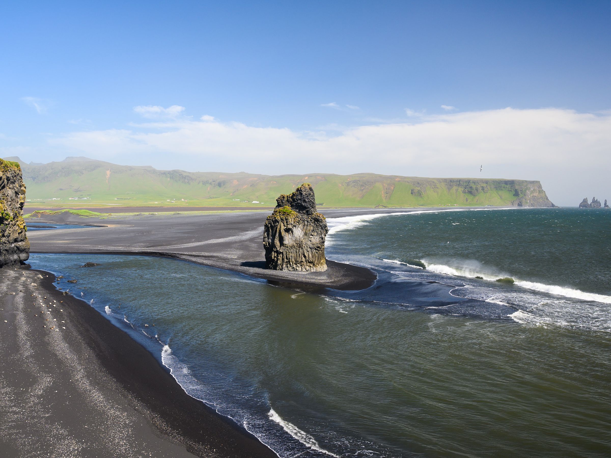 a black sand beach with a big rock in the middle on a sunny day