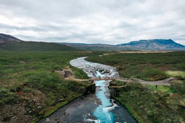 Turquoise Bruarfoss waterfall with a bridge over it, surrounded by green shrubs and distant mountains under a cloudy sky.