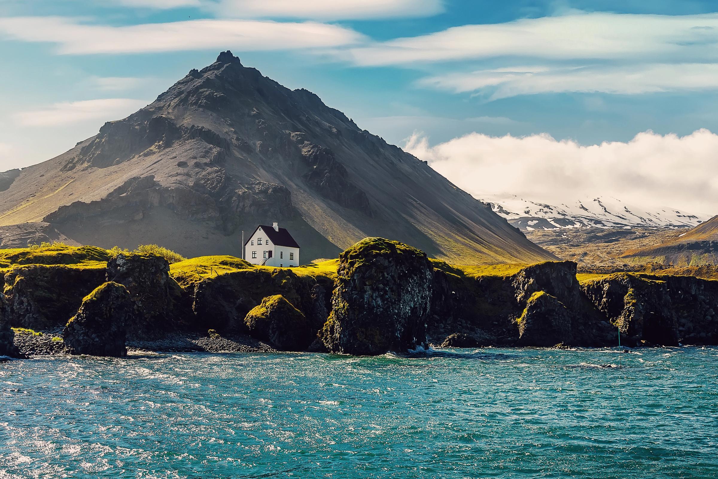 A white house sits on a green cliff overlooking a blue sea, with a large, dark mountain towering behind it.