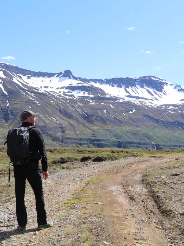 A hiker with a backpack stands on a dirt path, looking at snowy mountains under a clear sky.