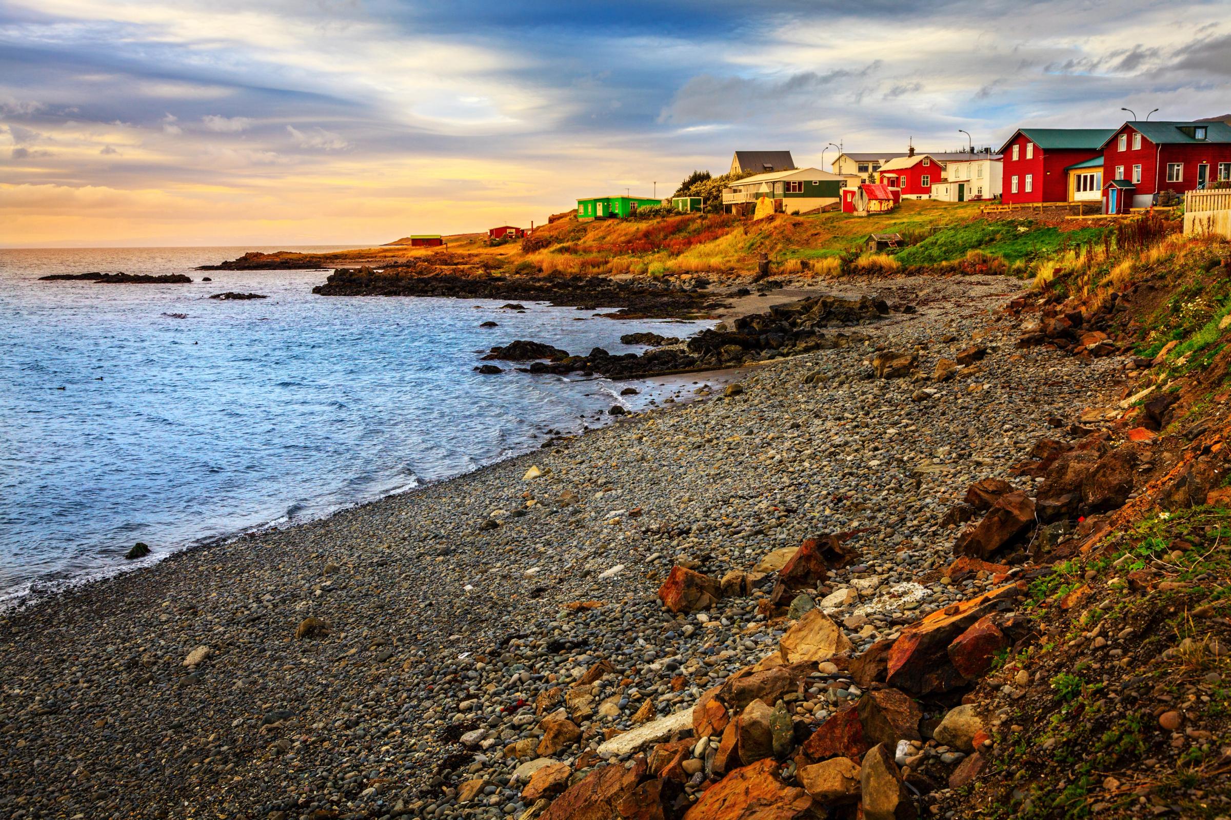 a row of red houses sit on a rocky shoreline next to the ocean, Hvammstangi