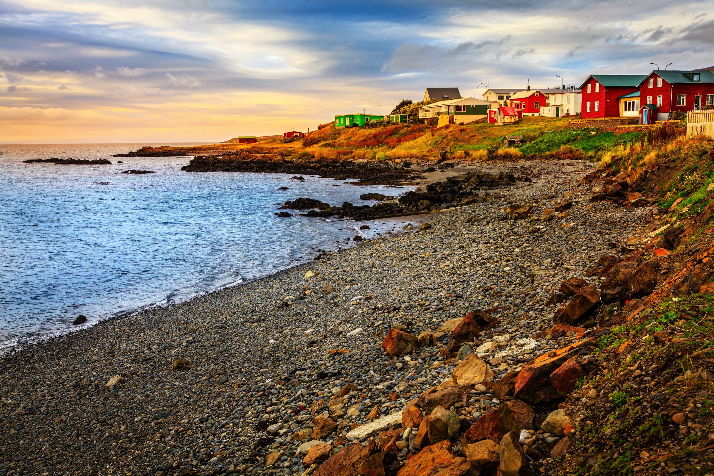 a row of red houses sit on a rocky shoreline next to the ocean, Hvammstangi
