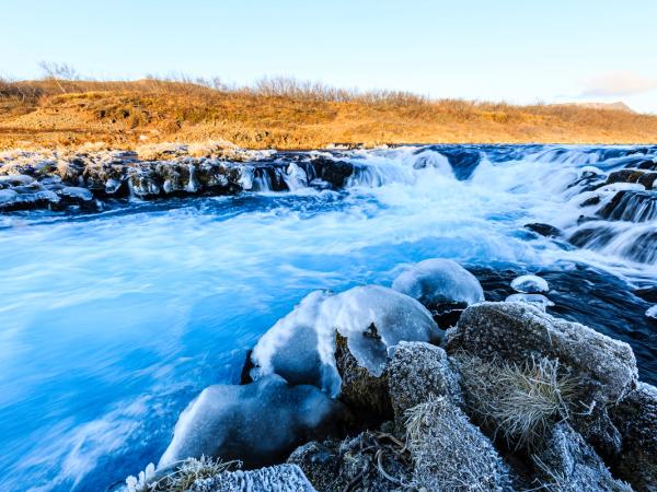 Bright blue river rapids flow over ice-covered rocks with golden hills in the background.
