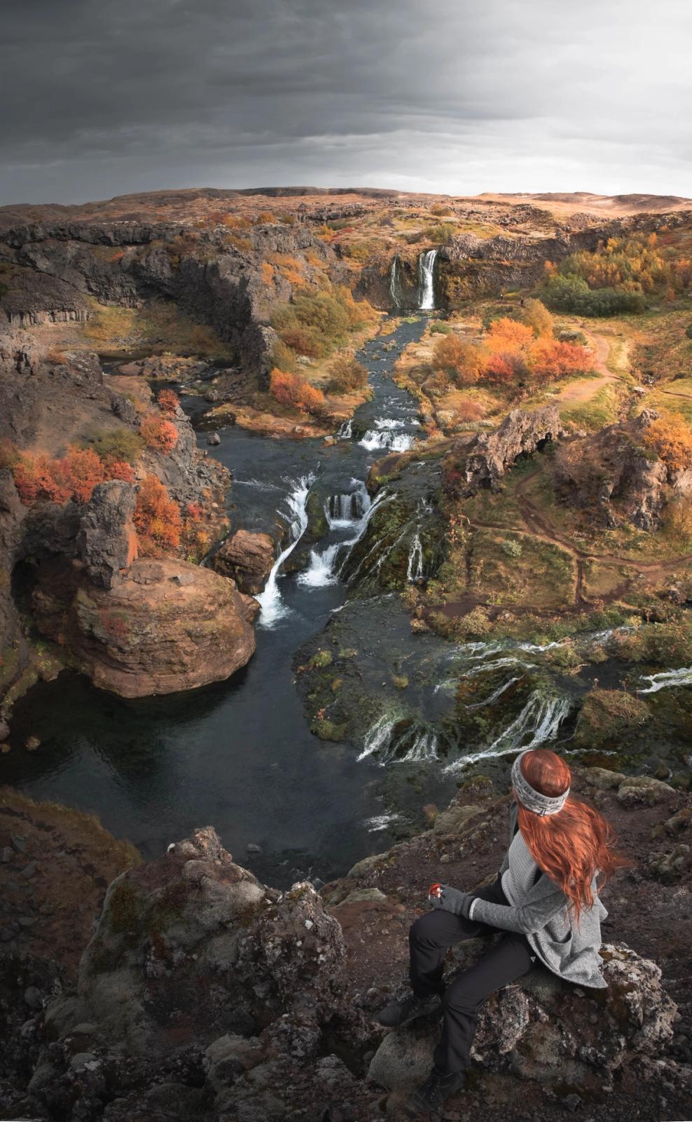 A red-haired woman sits on a rocky overlook, gazing at a river with multiple waterfalls winding through an autumnal canyon under an overcast sky.