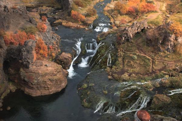 A red-haired woman sits on a rocky overlook, gazing at a river with multiple waterfalls winding through an autumnal canyon under an overcast sky.