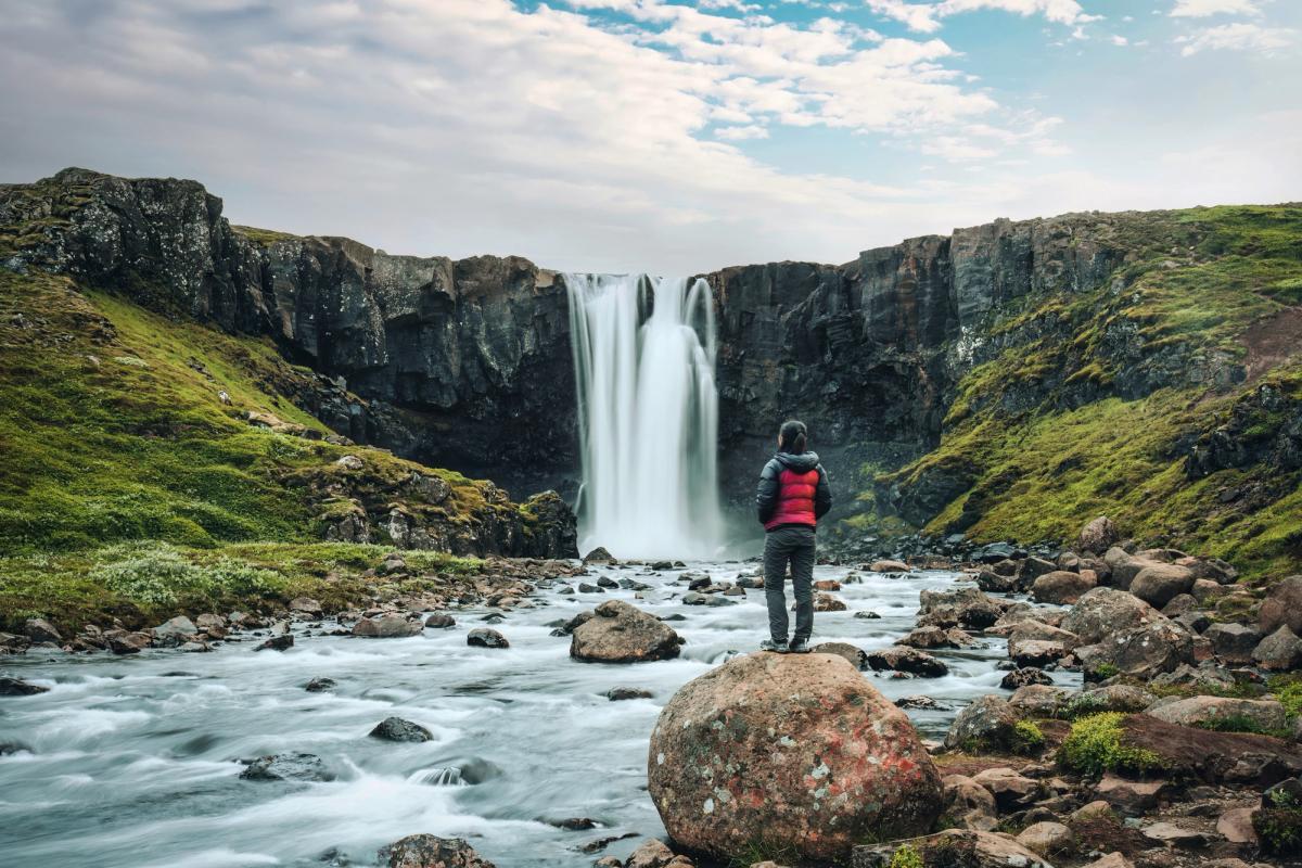 A person stands on a rock in a rocky river, facing a powerful waterfall surrounded by green cliffs.