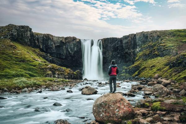 A person stands on a rock in a river, gazing at a large waterfall between grassy cliffs.
