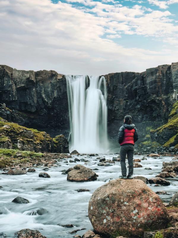A person stands on a rock in a rocky river, facing a powerful waterfall surrounded by green cliffs.
