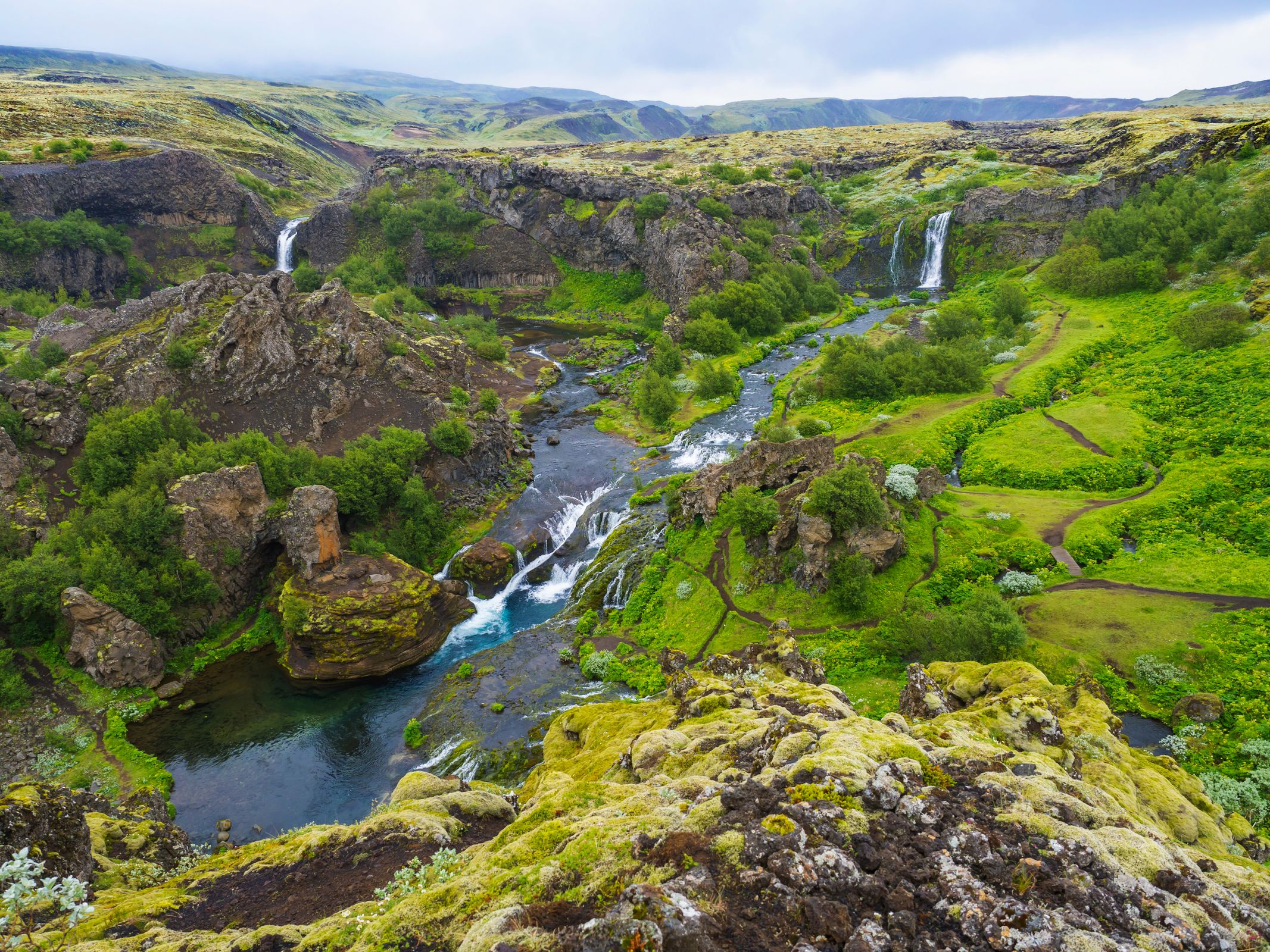 A lush green canyon with a river, two waterfalls, and winding paths.