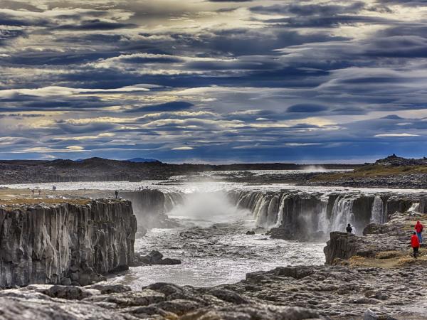 A powerful waterfall cascades through a wide, rocky canyon under a dramatic cloudy sky, with people observing from the barren banks.