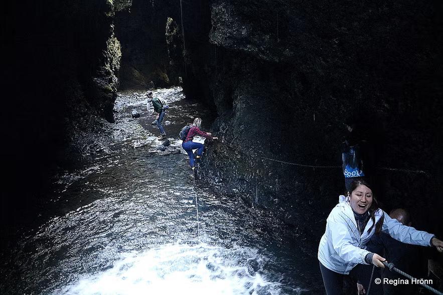 Un grupo de personas navega por un cañón oscuro y estrecho con un río que fluye, utilizando cuerdas a lo largo de las paredes rocosas.