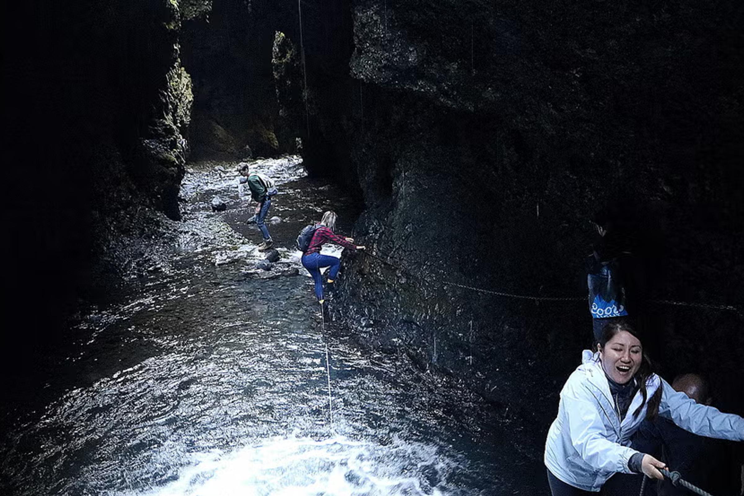 Un grupo de personas navega por un cañón oscuro y estrecho con un río que fluye, utilizando cuerdas a lo largo de las paredes rocosas.
