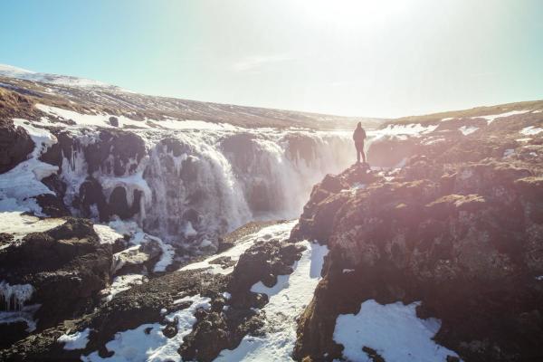 Person standing on a rocky, snowy cliff overlooking a powerful waterfall under a bright, sunny sky.