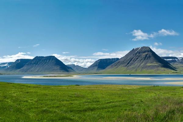 there is a house in the middle of a field next to a lake with mountains in the background .