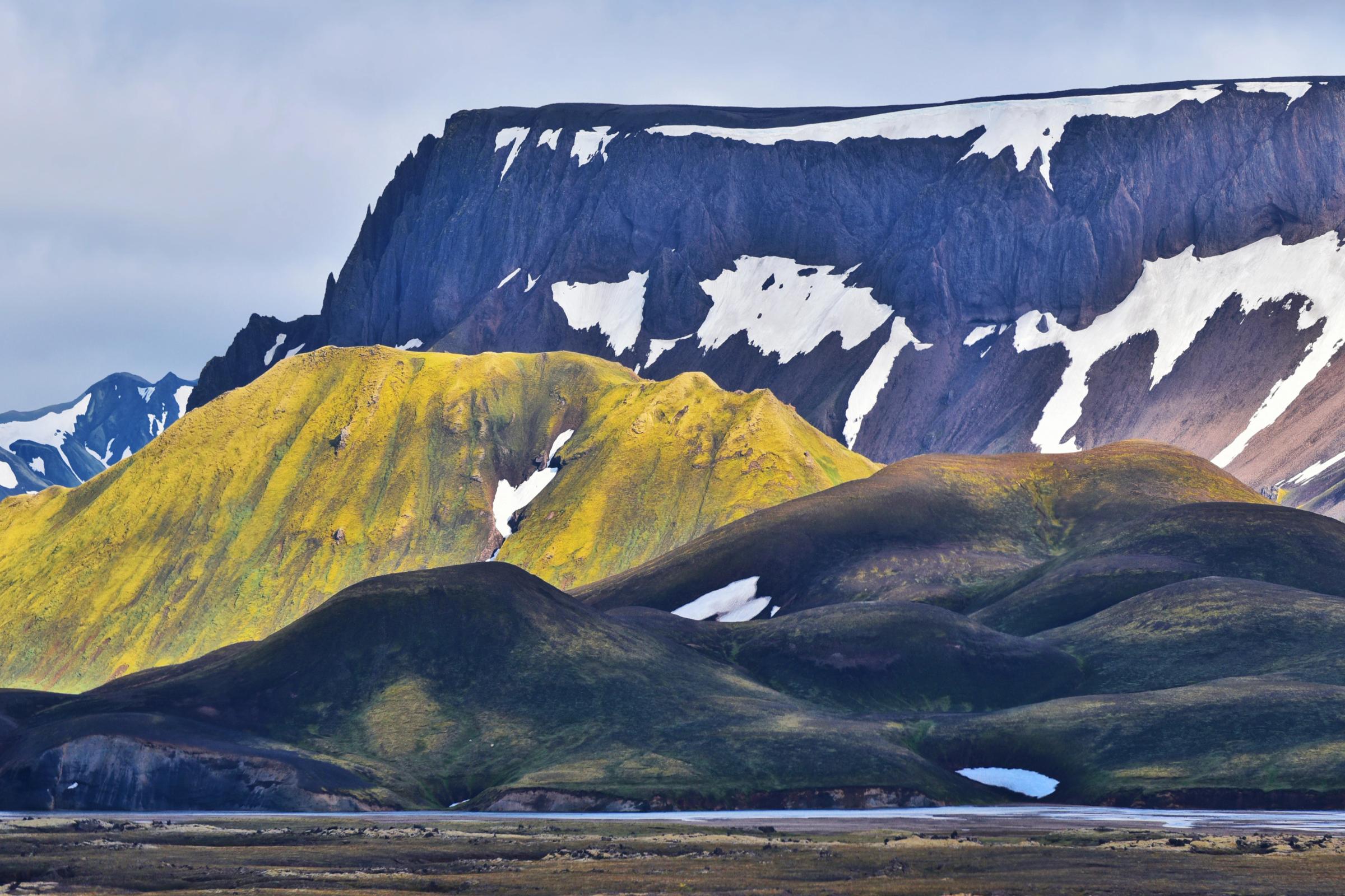 Panoramic view of a rugged landscape with a dark, snow-capped flat-topped mountain and vibrantly yellow-green and dark green hills.