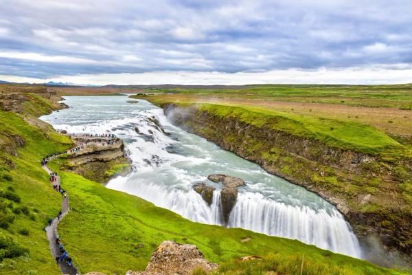 A powerful waterfall cascades into a canyon, surrounded by vibrant green landscapes, with a path full of tourists leading to a viewing area.