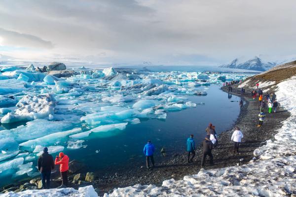 Tourists on a snowy, rocky shore observe a glacial lagoon filled with blue icebergs and snowy mountains.