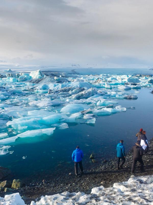 People walk along the snowy shore of a glacial lagoon filled with blue icebergs.