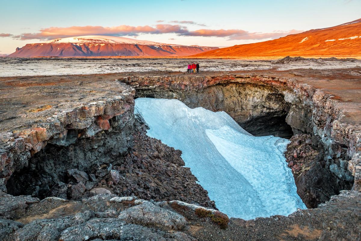 Una profunda cueva abierta en roca volcánica oscura, parcialmente llena de nieve, con personas en el borde y montañas bajo un cielo dorado.