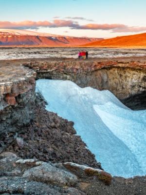 Una profunda cueva abierta en roca volcánica oscura, parcialmente llena de nieve, con personas en el borde y montañas bajo un cielo dorado.